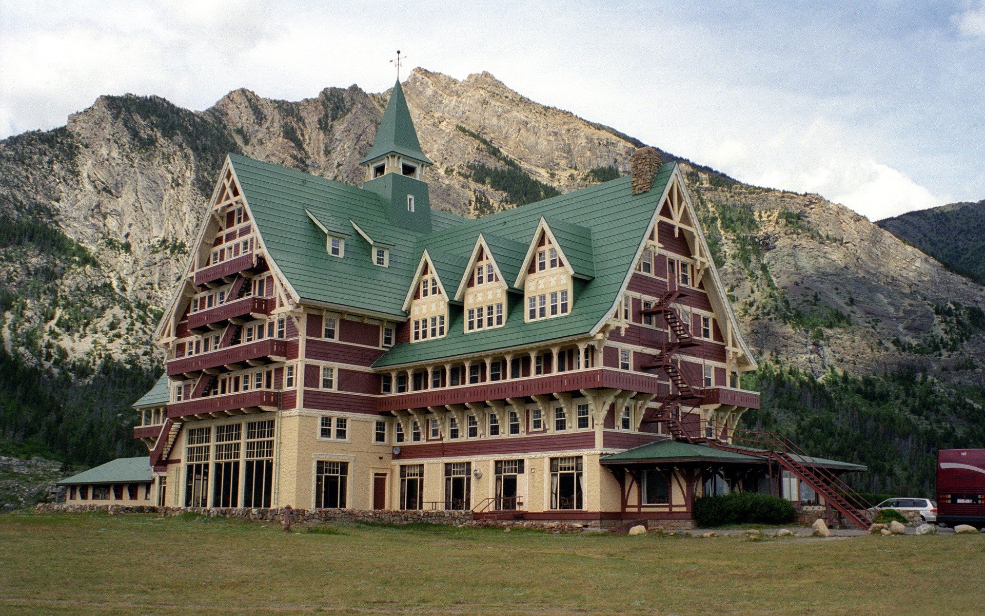 HD desktop wallpaper of a man-made historic hotel with green roofs set against rugged mountain peaks under a cloudy sky.