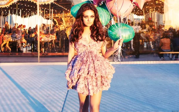 A glamorous Mila Kunis stands gracefully in a playful pink dress, holding colorful balloons, with a vibrant carnival scene in the background. HD wallpaper for a stylish desktop.