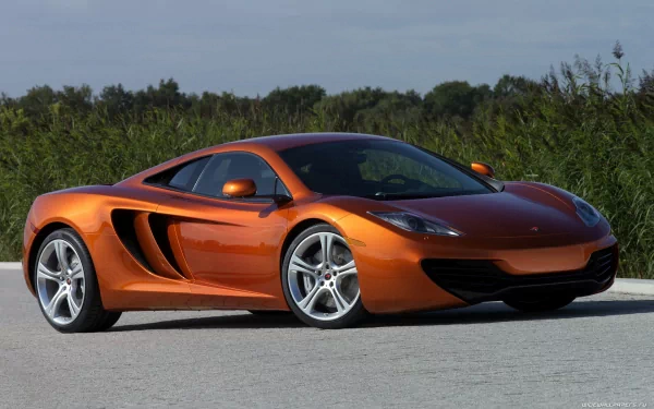 Orange McLaren MP4-12C supercar parked on a light pavement with greenery in the background, captured in high definition for PC desktop wallpaper.