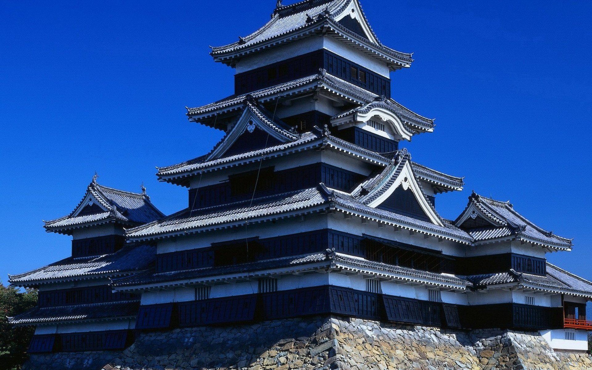 HD PC desktop wallpaper of Matsumoto Castle, Japan — a man-made, multi-tiered black castle on a stone base set against a vivid blue sky.