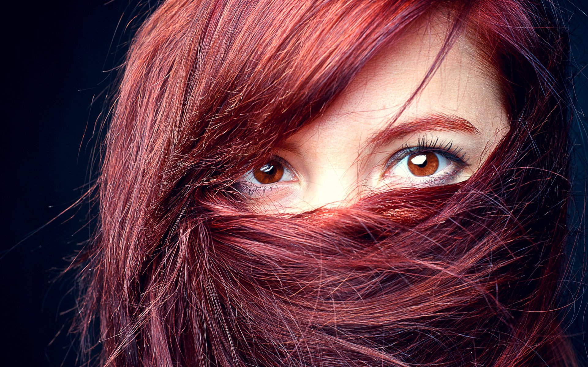 Close-up HD desktop wallpaper of a woman with vibrant red hair partially covering her face, highlighting her expressive brown eyes against a dark background.