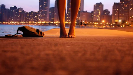 A close-up of a woman's bare feet on a sidewalk at sunset, with city skyscrapers and glowing lights in the background, creating a beautiful HD desktop wallpaper scene.