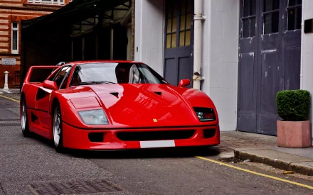 HD desktop wallpaper featuring a red Ferrari F40 parked on a street beside industrial-style buildings.
