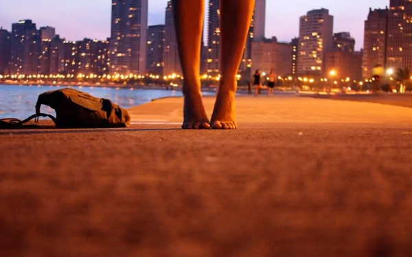 A close-up of a woman's bare feet on a sidewalk at sunset, with city skyscrapers and glowing lights in the background, creating a beautiful HD desktop wallpaper scene.