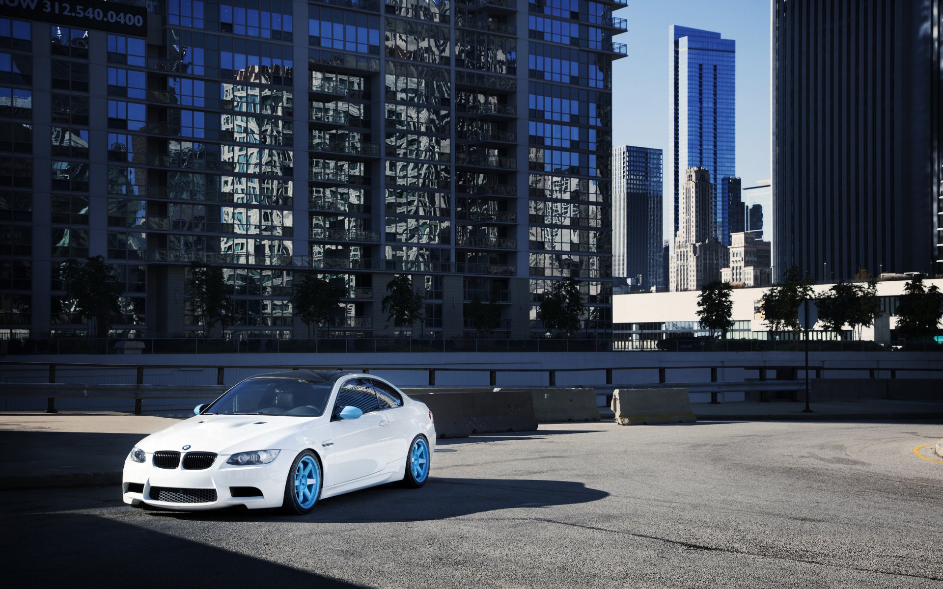 HD PC desktop wallpaper featuring a white BMW M3 with blue rims parked in an urban setting against tall modern buildings.