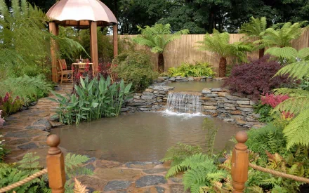 HD desktop wallpaper featuring a serene man-made garden with a small pond, cascading waterfall, lush greenery, and a gazebo in the background.