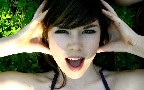 A close-up of a woman with long brown hair, smiling joyfully while lying on green grass. The vibrant colors create an engaging HD desktop wallpaper and background.