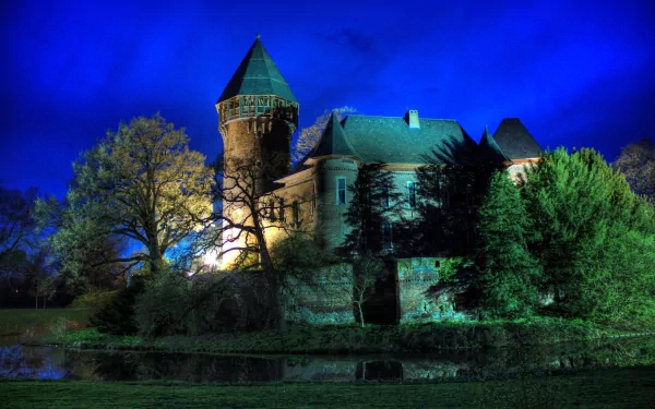HD desktop wallpaper of Krefeld Castle illuminated at night, surrounded by trees and reflected in calm water under a deep blue sky.