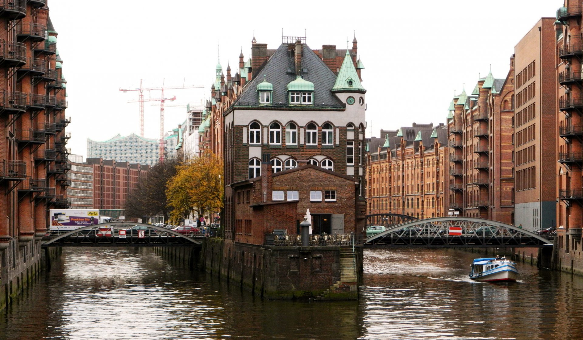HD desktop wallpaper showing Hamburg’s historic brick buildings and waterways with a boat cruising under a bridge in an urban man-made setting.