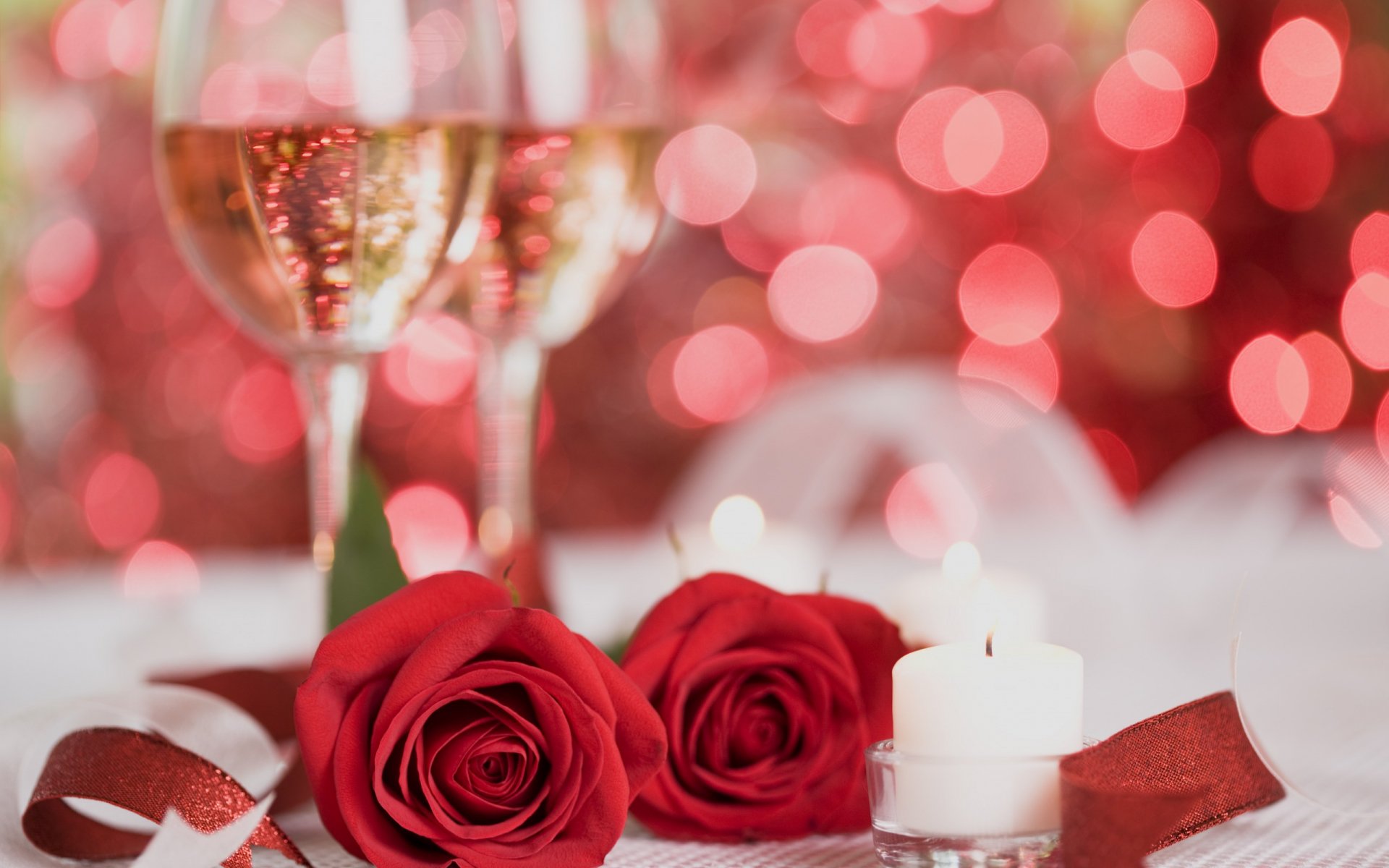 HD desktop wallpaper featuring two red roses, a lit candle, and two glasses of champagne against a sparkling red bokeh background celebrating Valentine's Day.
