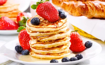 HD PC desktop wallpaper showcasing a stack of golden pancakes topped with fresh strawberries and blueberries on a white plate, with a croissant and more berries in the background.