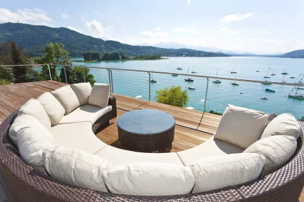 4K Ultra HD photography of a curved outdoor terrace sofa overlooking a serene bay with boats, mountains, and a partly cloudy sky in the background.
