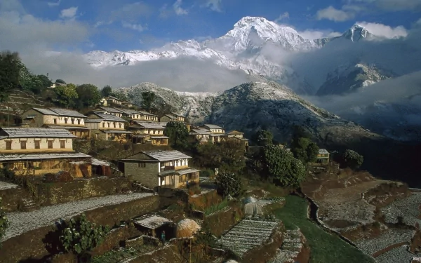 HD desktop wallpaper of a man-made town in Nepal nestled among green terraces with snow-capped mountains and a partly cloudy sky in the background.