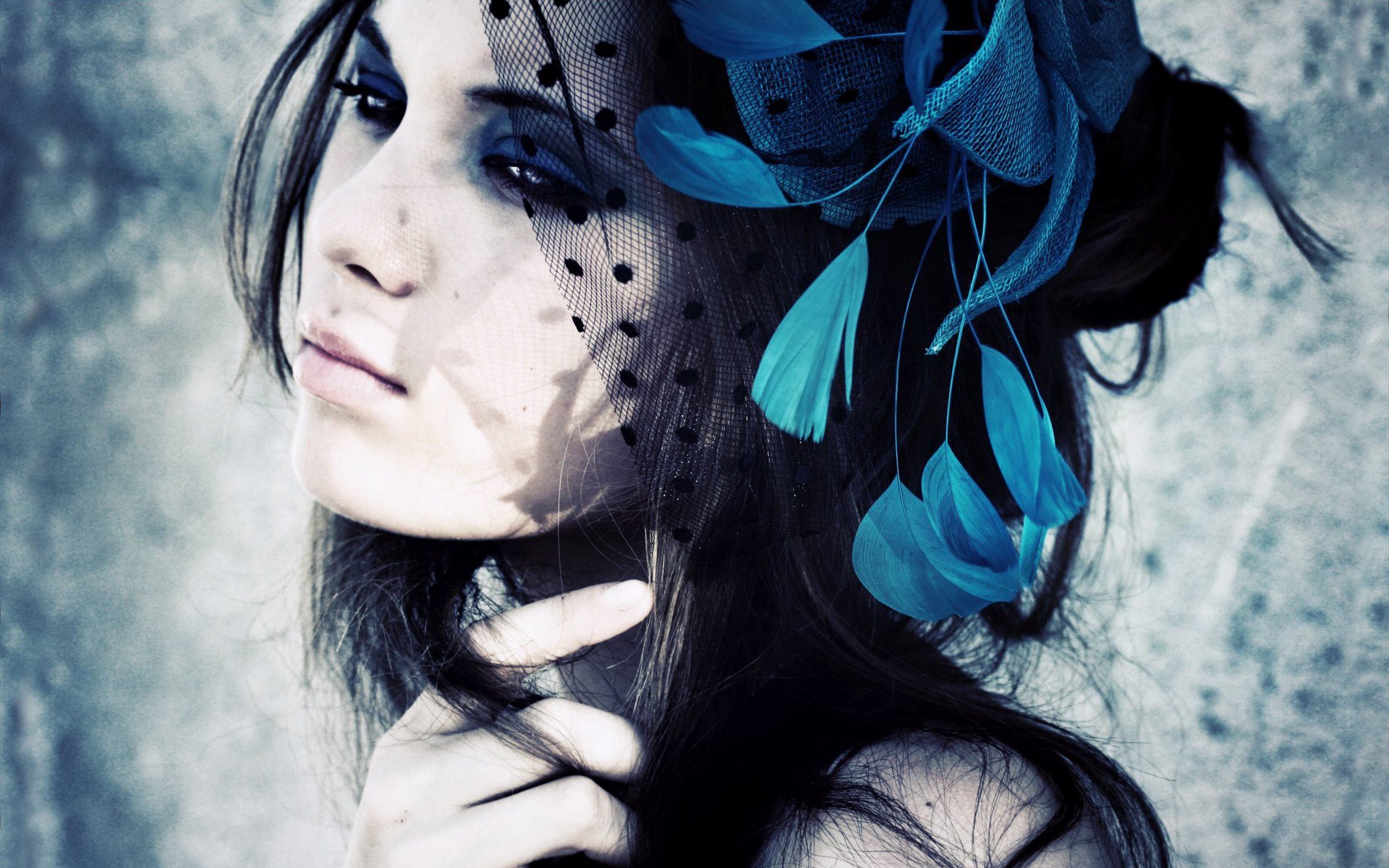 HD PC desktop wallpaper of a woman model with a dotted black veil and blue feathered headpiece, dark eye makeup, hand at chin against a moody textured background.