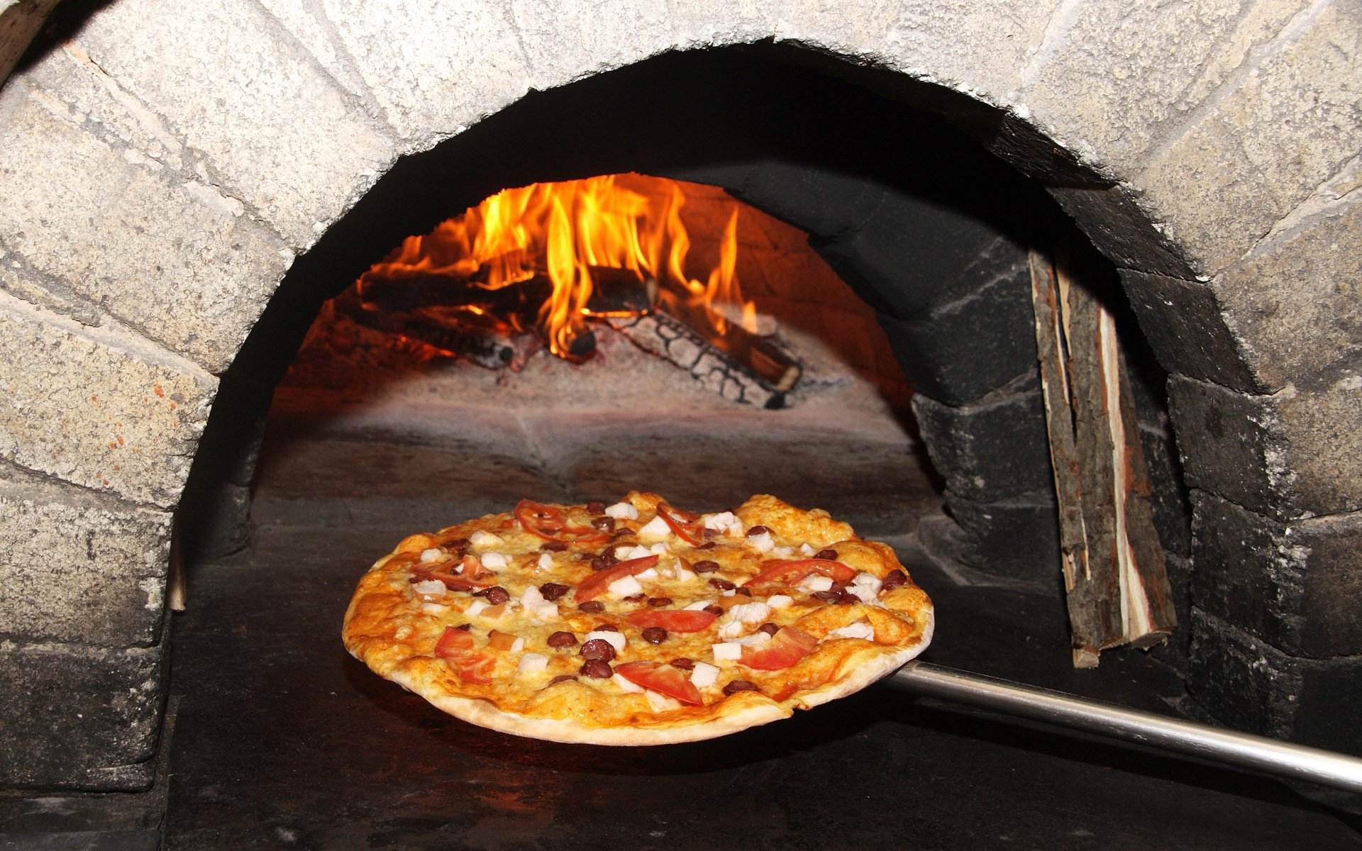 HD PC desktop wallpaper showing a pizza on a peel being slid into a wood-fired stone oven, flames glowing as they crisp the crust.