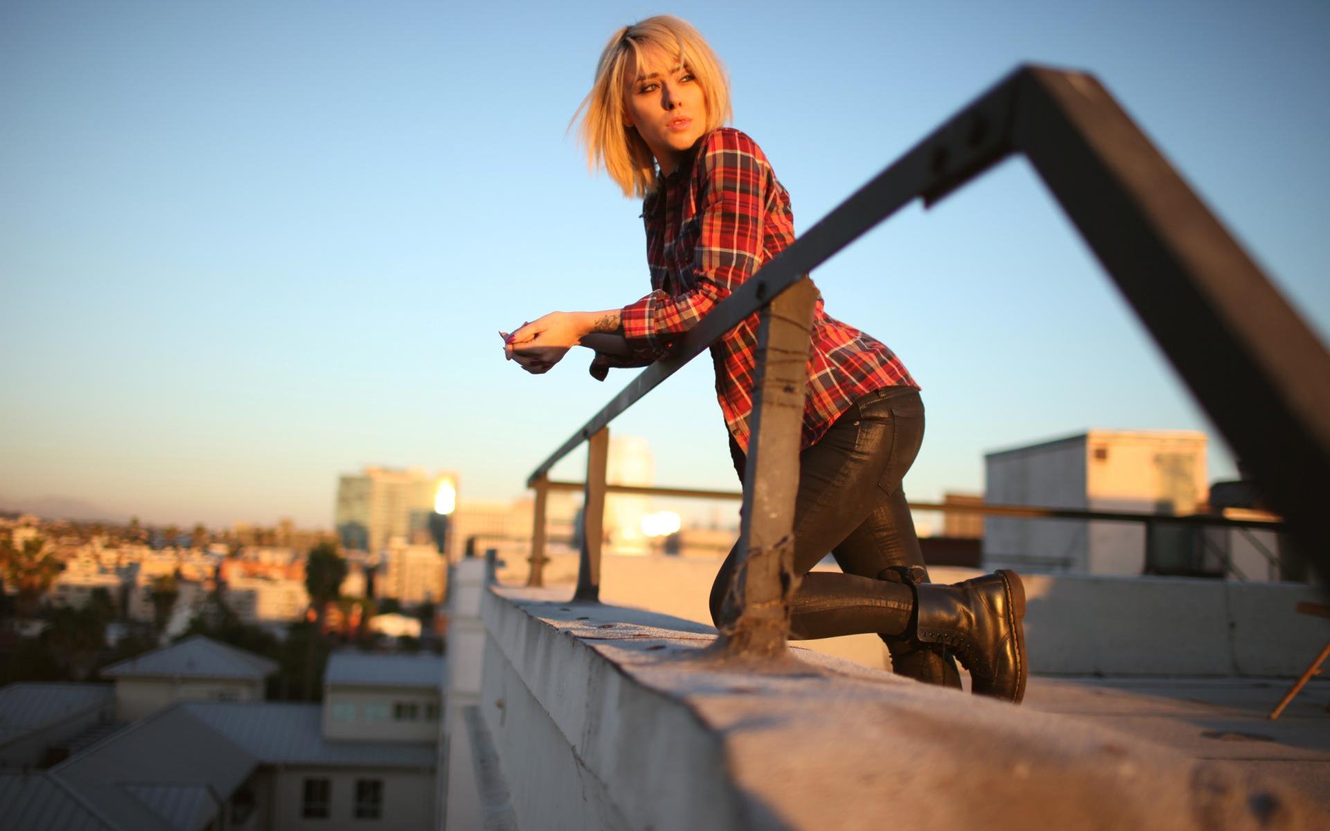 HD PC desktop wallpaper featuring a woman model in a red plaid shirt and black pants posing on a rooftop railing at sunset with a cityscape background.