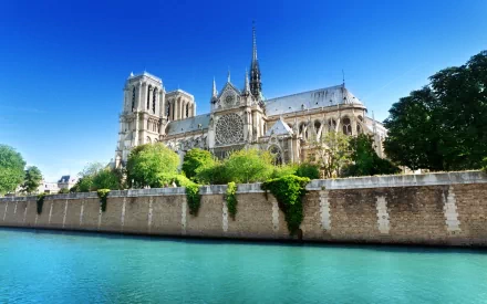 HD desktop wallpaper showing the iconic Notre-Dame de Paris cathedral under a clear blue sky, with the Seine River and lush greenery in the foreground.