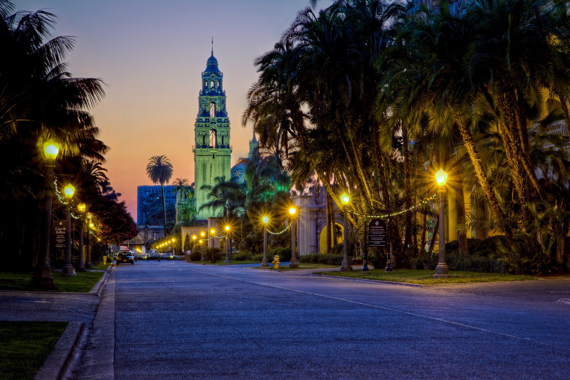 2K Quad HD PC desktop wallpaper and background of a tree-lined road at dusk leading to a lit man-made bell tower, palm trees and streetlights glowing under a twilight sky.