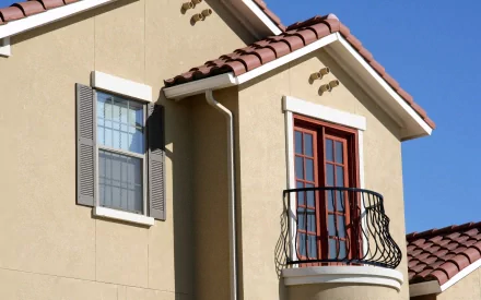 2K Quad HD PC desktop wallpaper showing a man-made villa facade: stucco walls, red-tiled roof, shuttered window and wrought-iron balcony under a clear blue sky.