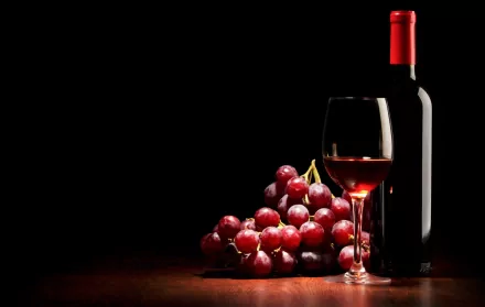 4K Ultra HD wallpaper featuring a bottle of red wine, a glass filled with wine, and a bunch of red grapes on a dark wooden surface against a black background.