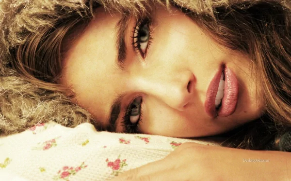 A close-up image of a woman with soft features, featuring long eyelashes and full lips, lying on a floral-patterned surface, creating an intimate and serene ambiance.