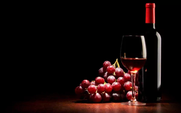 4K Ultra HD wallpaper featuring a bottle of red wine, a glass filled with wine, and a bunch of red grapes on a dark wooden surface against a black background.