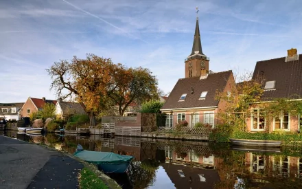 HD PC desktop wallpaper and background: man-made village canal with quaint brick houses, a church steeple, trees and moored boats reflecting in calm water under a blue sky.