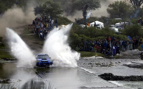 HD PC desktop wallpaper featuring a Subaru Impreza rally car splashing through a water crossing with spectators and trees in the background.