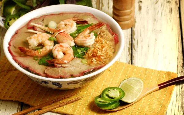 HD PC desktop wallpaper showing a bowl of shrimp soup garnished with herbs, sliced jalapeños, and lime on a rustic wooden table.