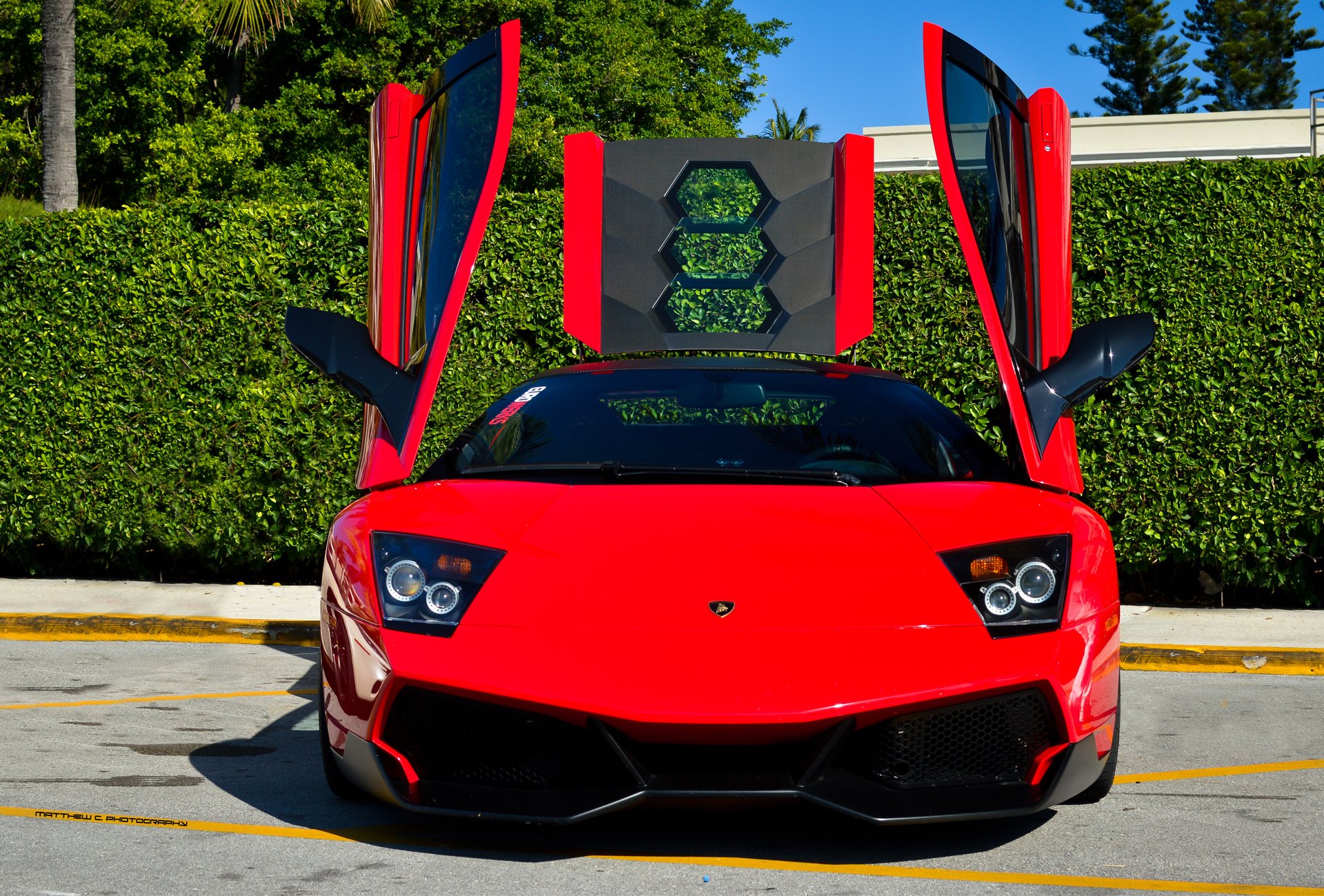 HD desktop wallpaper featuring a red Lamborghini Murcielago with its scissor doors open, set against a backdrop of greenery and clear blue sky.