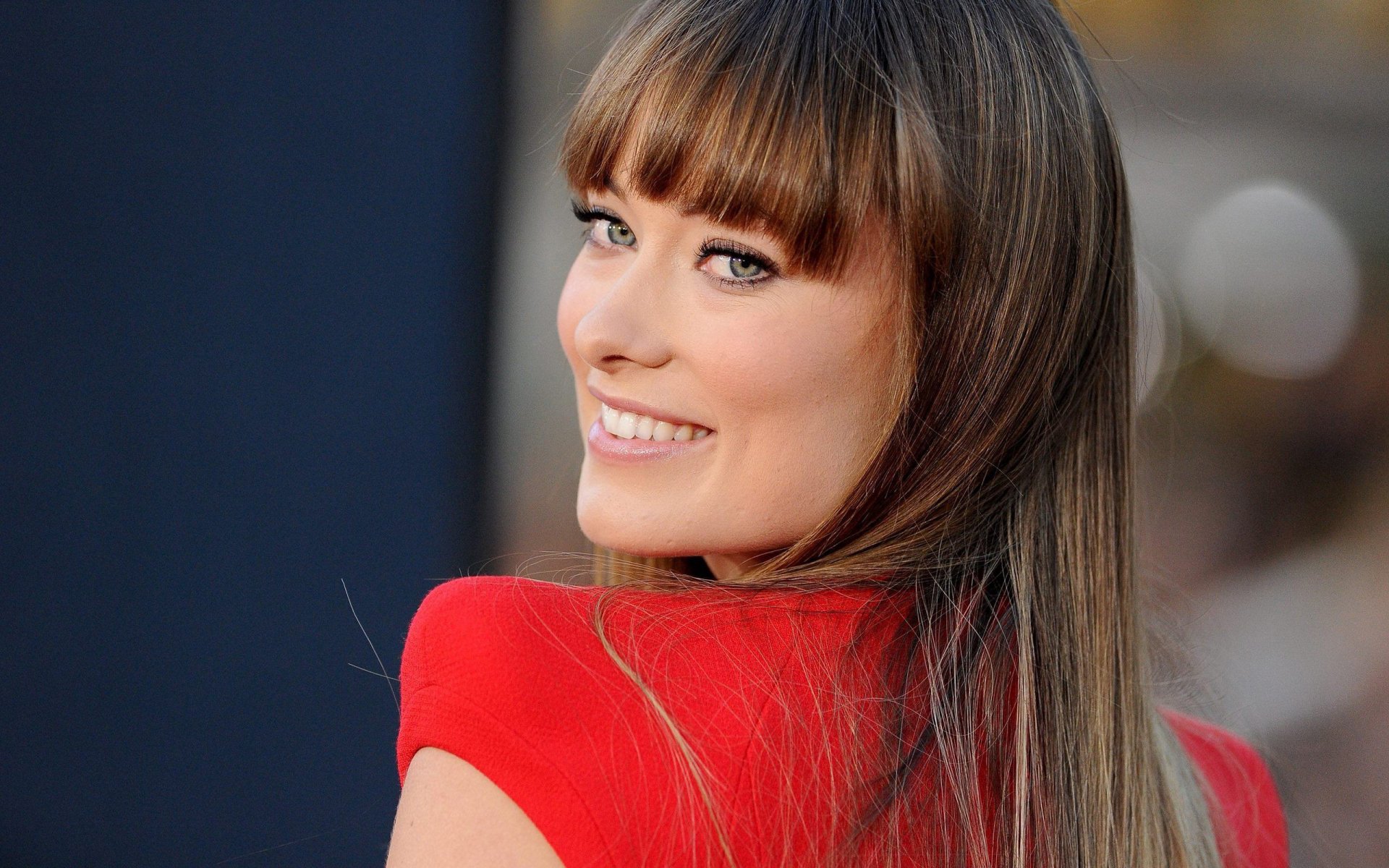 HD PC desktop wallpaper featuring celebrity Olivia Wilde smiling and looking over her shoulder, wearing a red outfit with a blurred background.