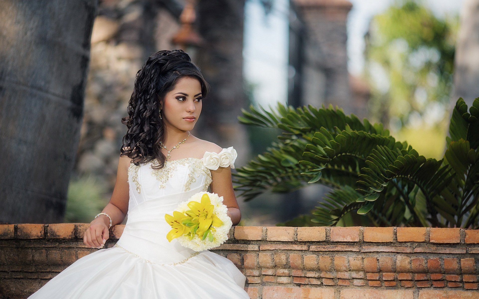 HD PC desktop wallpaper and background: bride in a white gown holding a yellow bouquet, seated on a brick wall with lush ferns and blurred trees behind her.
