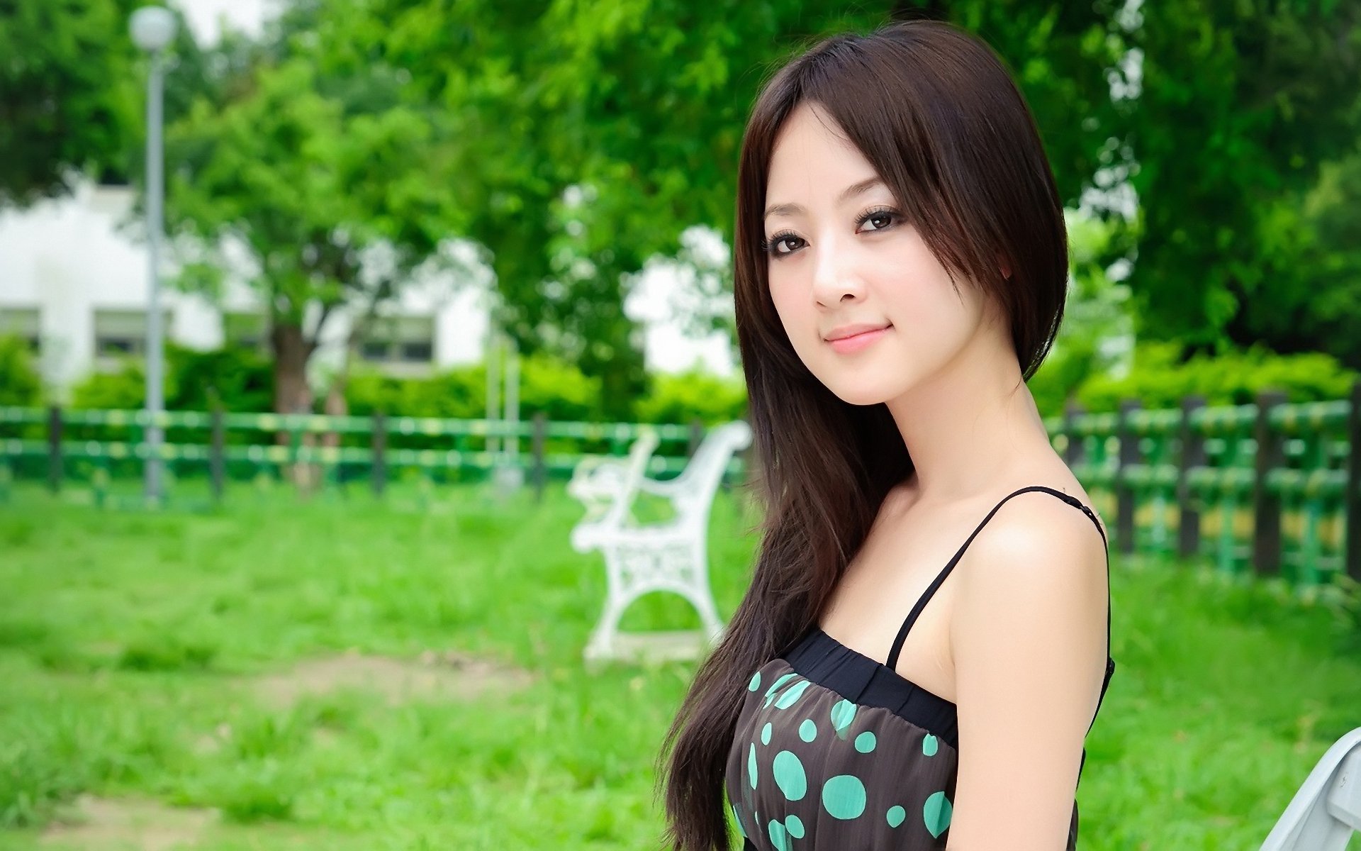 A model with long hair smiles softly while seated in a green park, surrounded by trees and empty benches. This image serves as an HD desktop wallpaper and background.