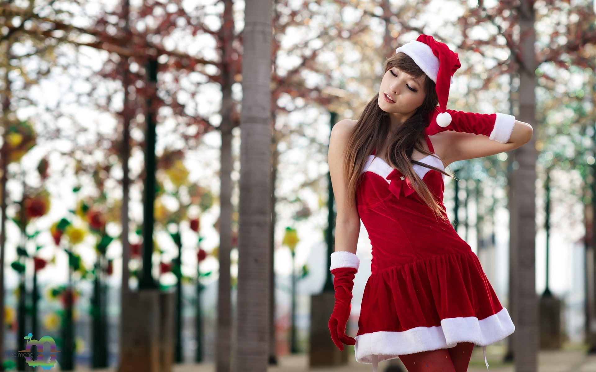 A brunette Asian woman in a festive Santa hat and red dress poses outdoors, surrounded by trees and colorful decorations. HD desktop wallpaper and background.