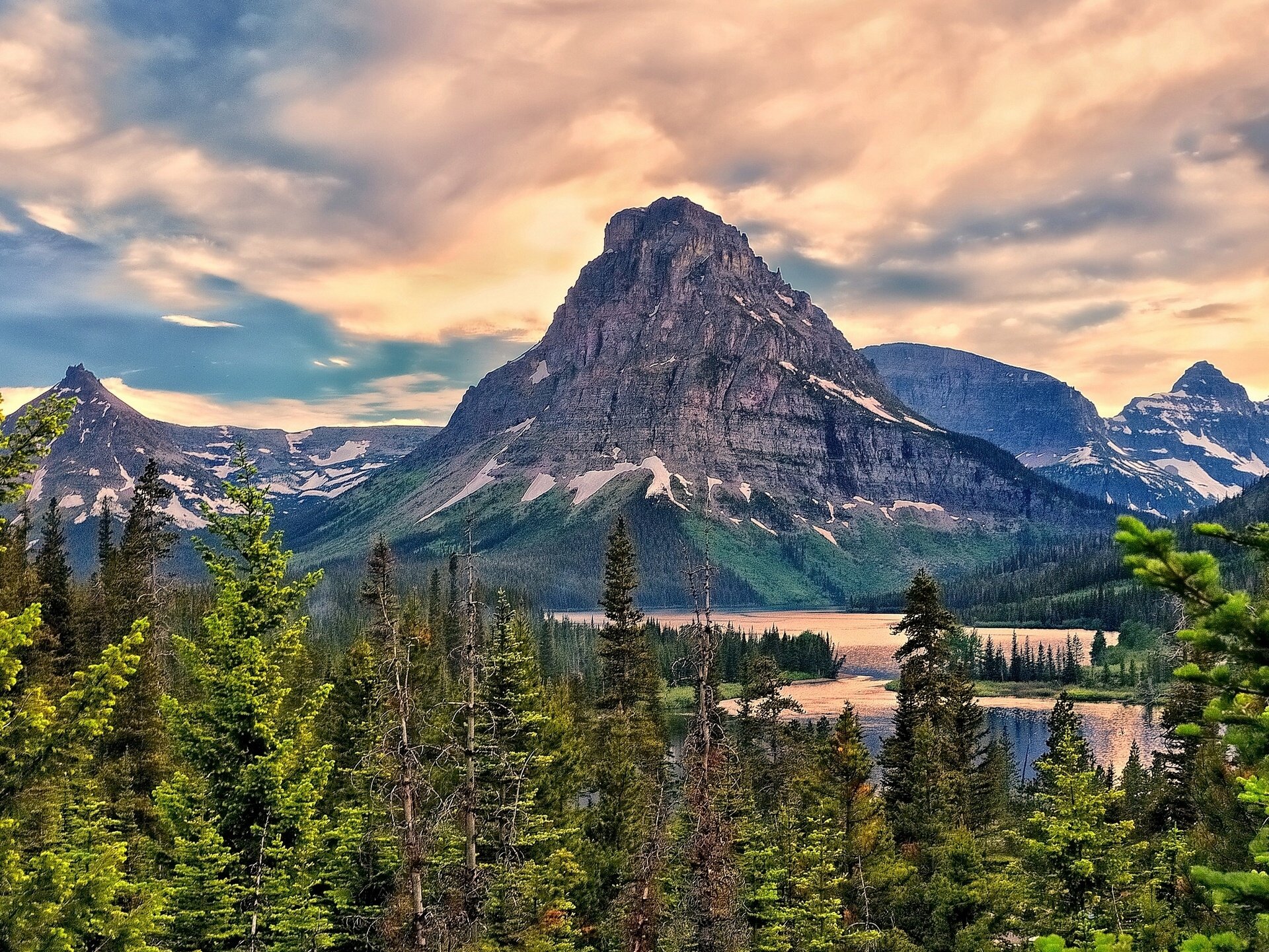 HD desktop wallpaper showcasing Sinopah Mountain towering over a serene lake surrounded by lush forests in Glacier National Park under a vibrant sky.