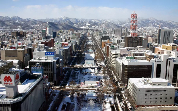 HD desktop wallpaper of Sapporo, Hokkaido — snow-dusted man-made cityscape with a central tree-lined avenue, urban towers and distant mountains under a clear sky.