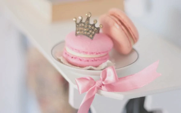 A charming HD wallpaper featuring pink macarons on a delicate plate, adorned with a crown and ribbon, capturing the elegance of dessert presentation.