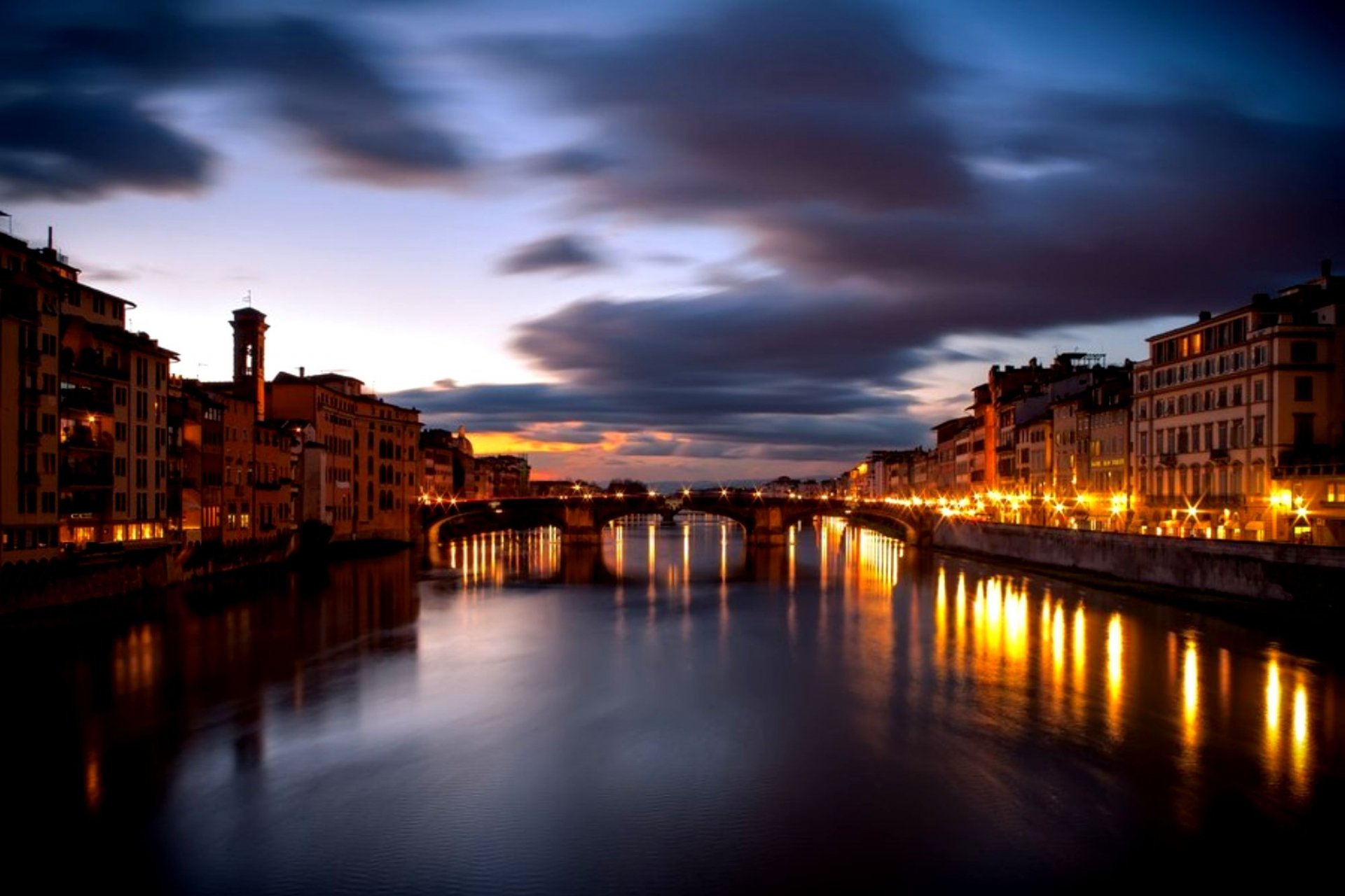 HD desktop wallpaper showcasing Florence's man-made architecture along a river at dusk, with illuminated buildings reflecting on calm water under a dramatic sky.