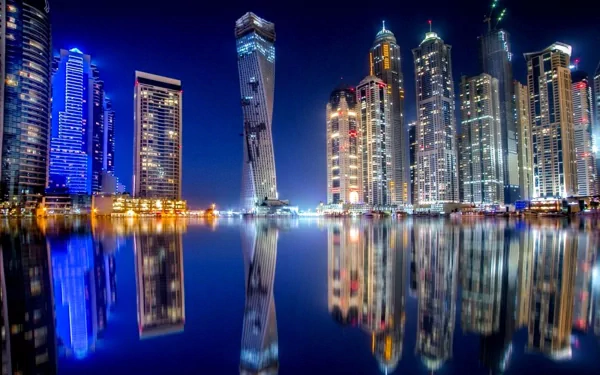 A stunning night view of Dubai's skyline, showcasing iconic skyscrapers illuminated against the dark sky, reflected in the calm water below. An impressive man-made urban landscape.