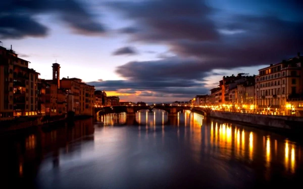 HD desktop wallpaper showcasing Florence's man-made architecture along a river at dusk, with illuminated buildings reflecting on calm water under a dramatic sky.
