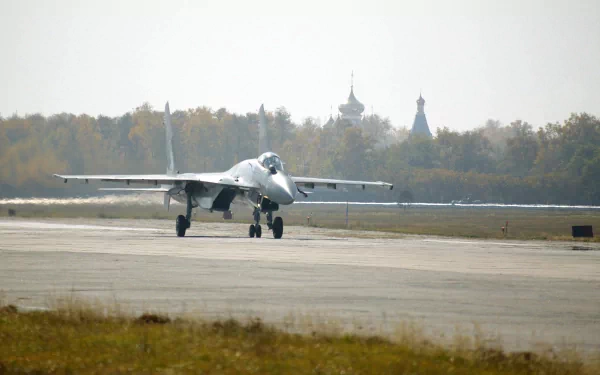 HD PC desktop wallpaper featuring a Sukhoi Su-35 military jet taxiing on a runway with trees and distant buildings in the background.