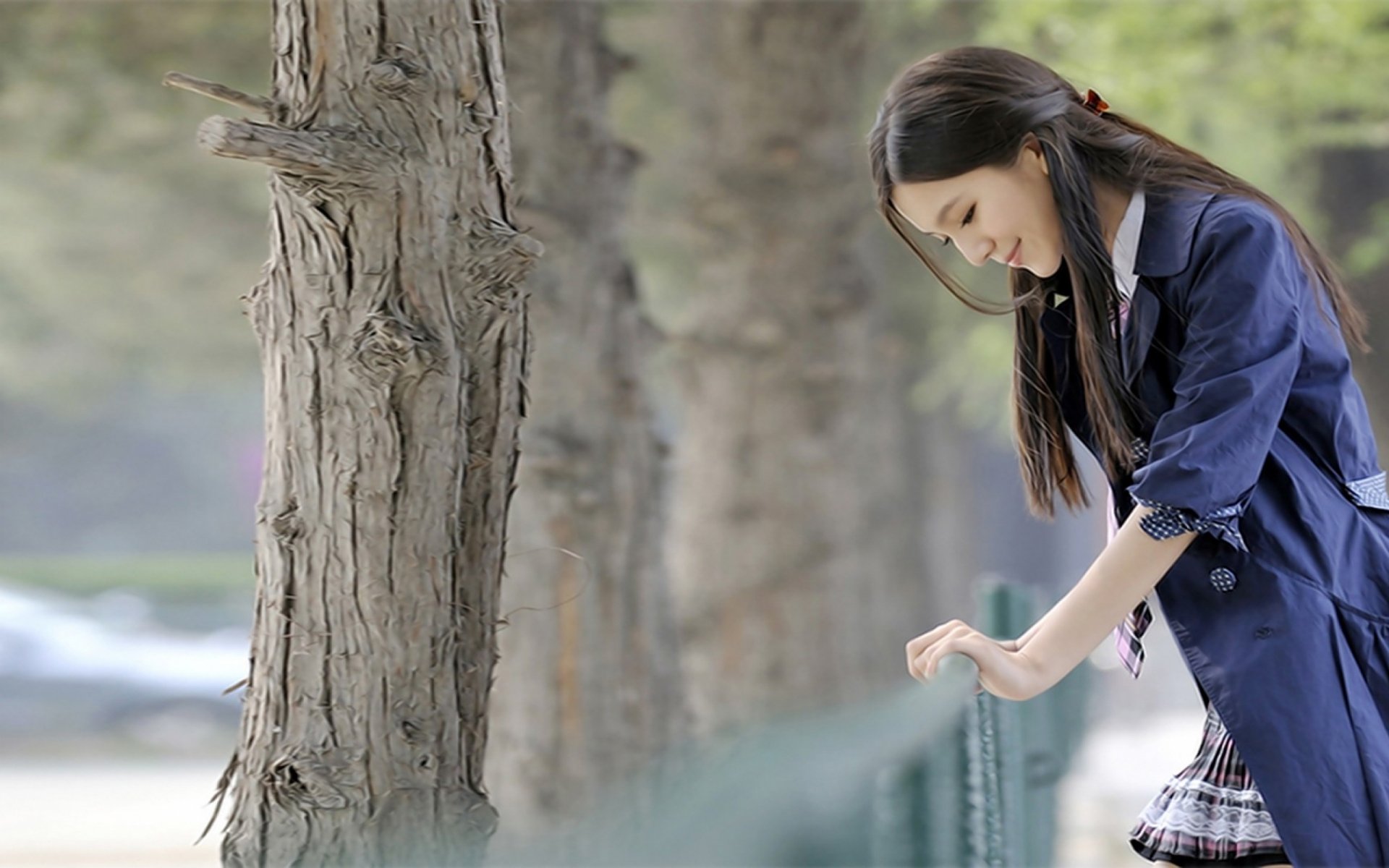 HD desktop wallpaper of an Asian woman standing near trees, looking down thoughtfully with a serene expression.