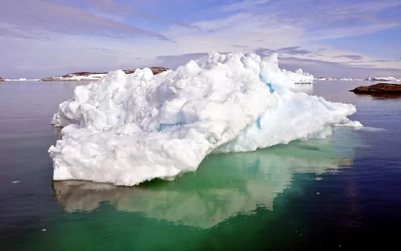 HD desktop wallpaper of a large iceberg floating in calm, clear waters under a partly cloudy sky, showcasing the serene beauty of nature.