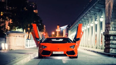 HD wallpaper featuring a striking orange Lamborghini Aventador with its doors open, parked at night on a city street illuminated by streetlights.