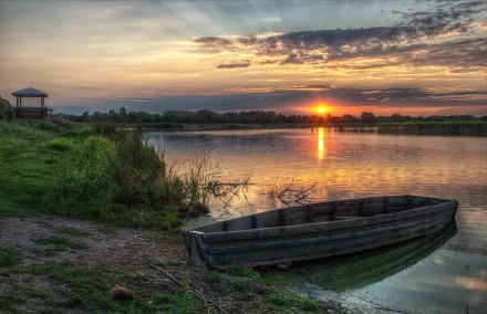 HD desktop wallpaper of a wooden boat resting on a calm lakeshore at sunset, with vibrant skies reflecting on the water and a small pavilion in the distance.
