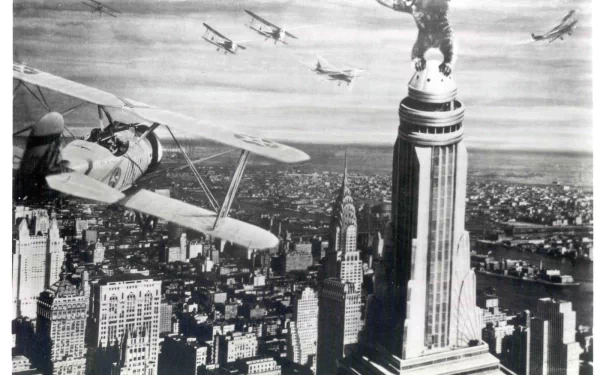 A dramatic scene from King Kong (1933) featuring the giant ape atop the Empire State Building, with biplanes flying around in the skyline of a bustling city. HD desktop wallpaper.