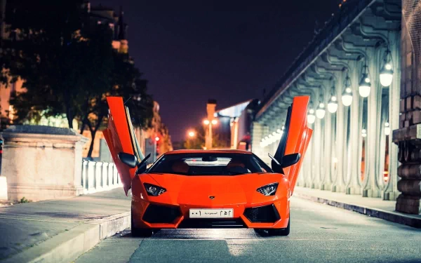 HD wallpaper featuring a striking orange Lamborghini Aventador with its doors open, parked at night on a city street illuminated by streetlights.