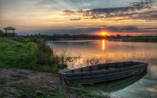HD desktop wallpaper of a wooden boat resting on a calm lakeshore at sunset, with vibrant skies reflecting on the water and a small pavilion in the distance.