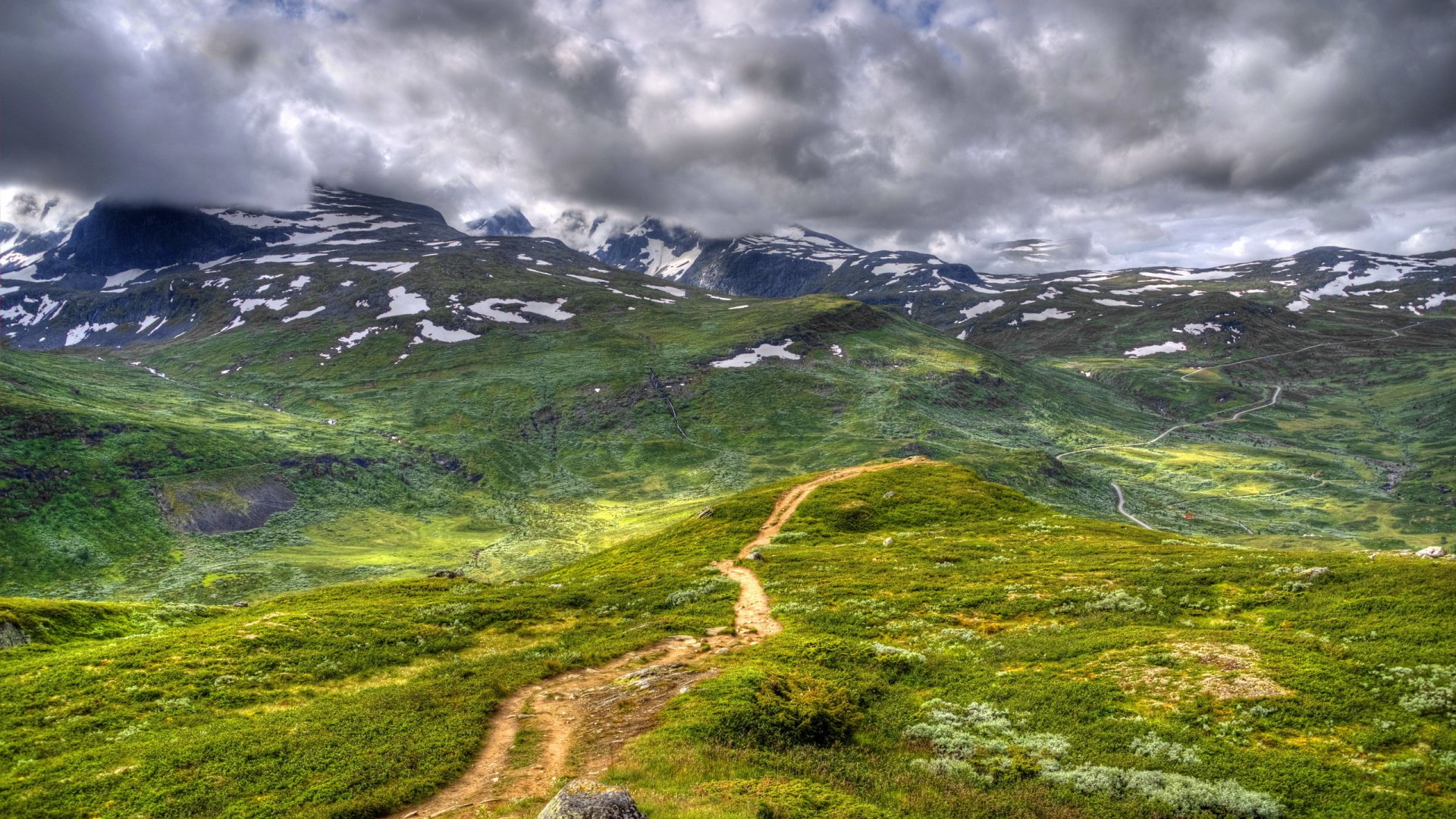 HD nature landscape showcasing a winding trail through lush green hills with snow-capped mountains and dramatic cloudy skies, designed as a PC desktop wallpaper.