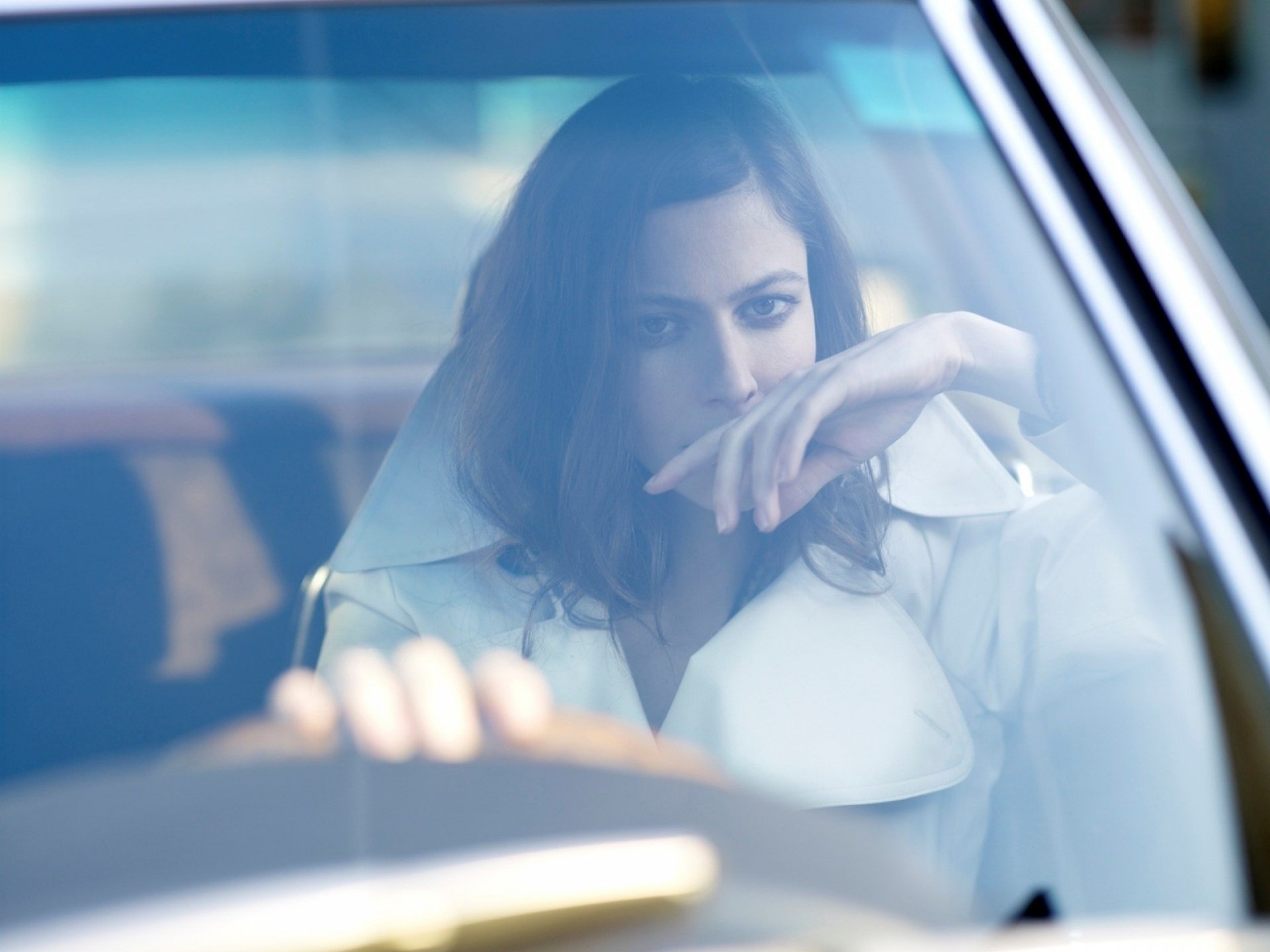 Woman in a white coat seen through a car windshield, hand near her face, soft cinematic blue lighting — 2K Quad HD PC desktop wallpaper and background.
